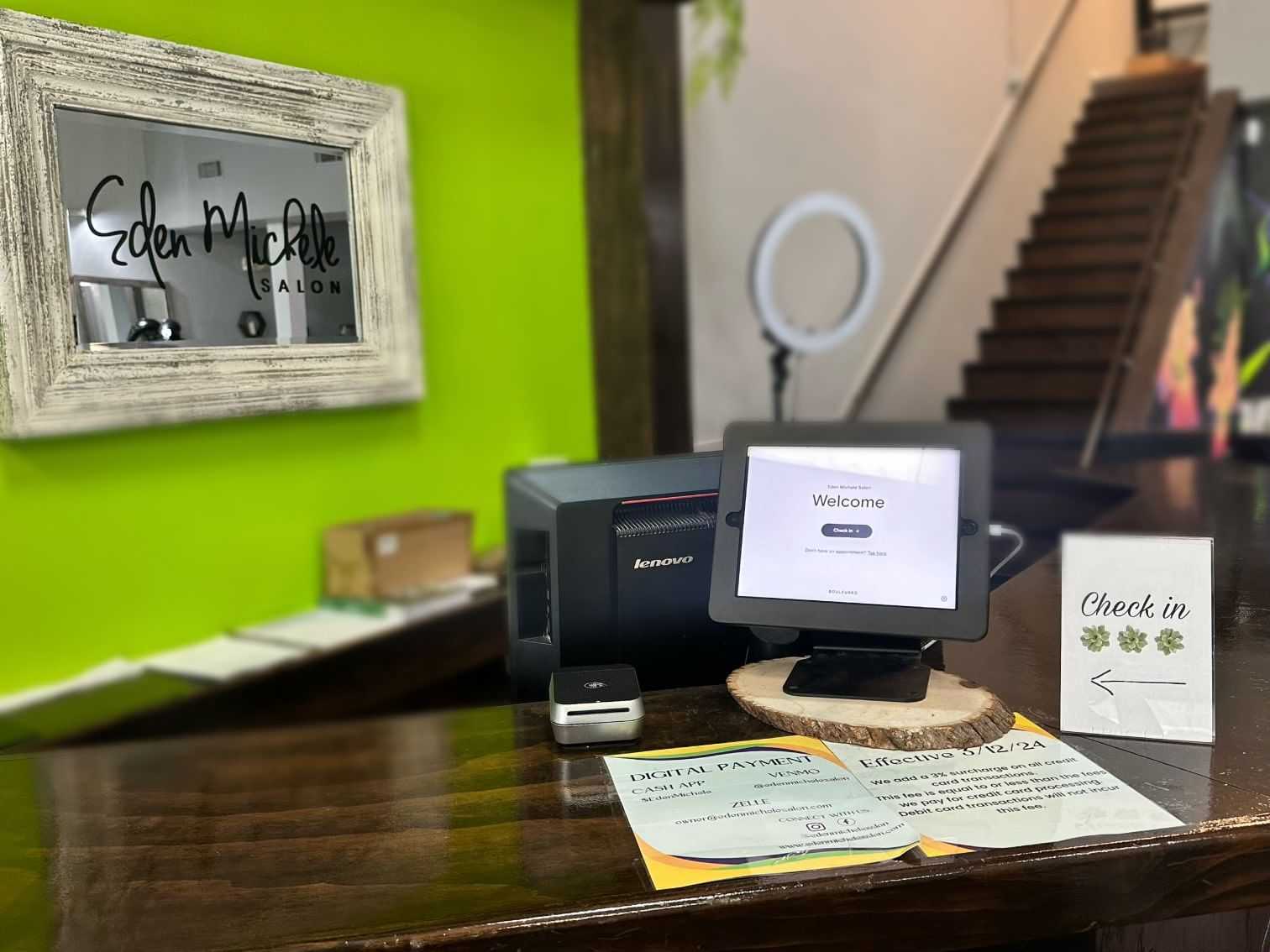 Reception desk with a computer, check-in tablet, and "Eden Michele Salon" sign on a green wall.